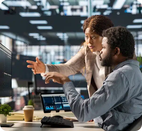 Two colleagues work together at a computer in a modern office. One person sits and types while the other leans over, pointing at the screen, both focused on data displayed on multiple monitors.