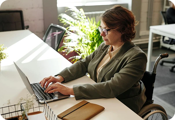 A woman in a wheelchair, wearing glasses and a blazer, types on a laptop at a desk in a bright office. A notebook and plant are on the desk, and sunlight streams through the window behind her.