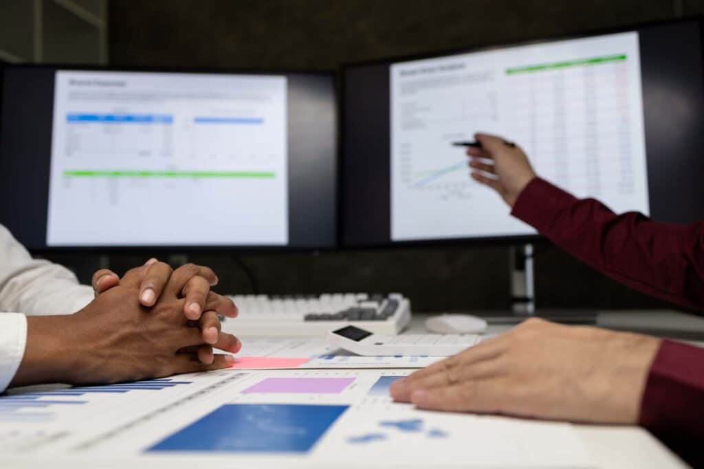 Two people sit at a desk covered with charts and documents, discussing wip reporting as one points at data on a large monitor. Two screens in the background display graphs and spreadsheets, highlighting financial health for construction companies.