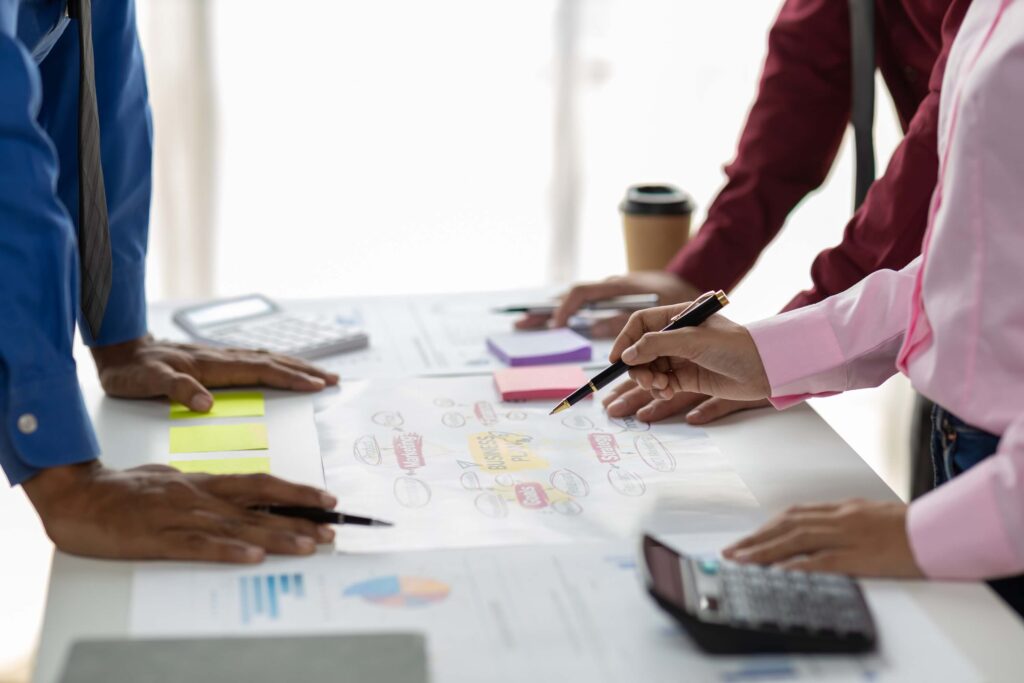 Three people stand around a table, holding pens and pointing at documents and a large paper with diagrams, focusing on project financial management. A calculator, sticky notes, and coffee cup suggest a collaborative work meeting.