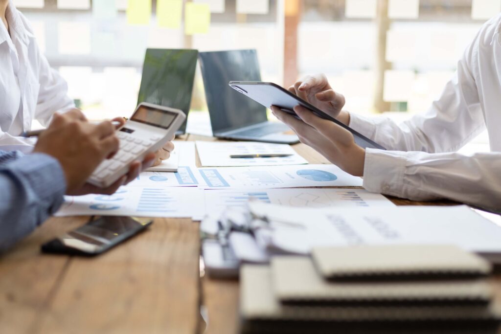 Three people sit at a wooden table with documents, charts, and devices, discussing data and analyzing business reports in a bright office, as they address overdue payments and work to improve their invoicing process.