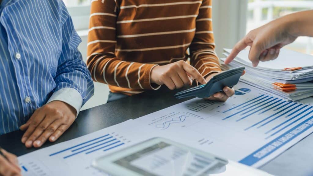 Three people at a table reviewing financial charts and graphs. One uses a calculator while another points at documents, demonstrating the account reconciliation process. Stacks of paperwork and a tablet are also visible on the table.