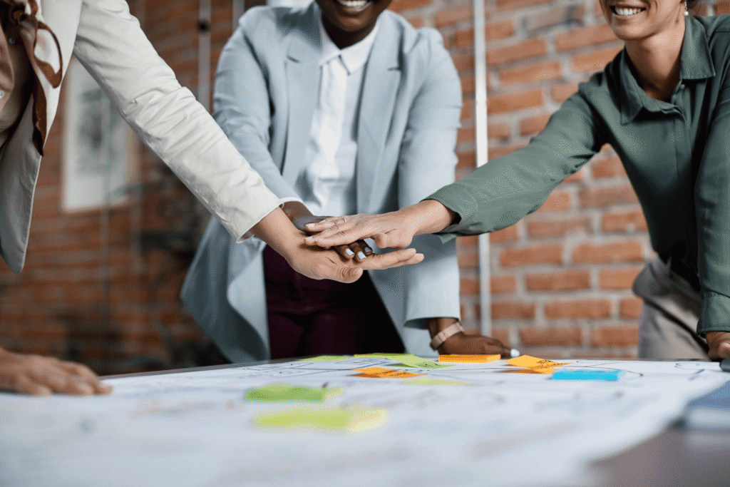 Three people stand around a table covered with papers and sticky notes, placing their hands together in a gesture of teamwork—demonstrating strong project management skills with a brick wall in the background.