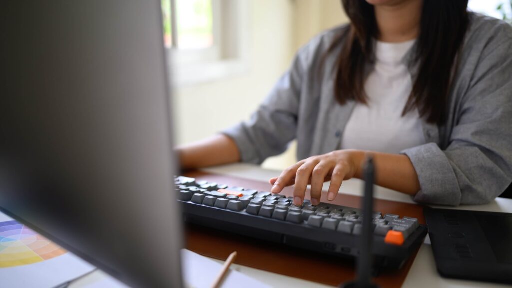 A person in a gray shirt types on a desktop computer keyboard at a desk, focusing on their hands as they address an invoicing problem. A monitor and some office supplies are visible in the foreground, framing the workspace.