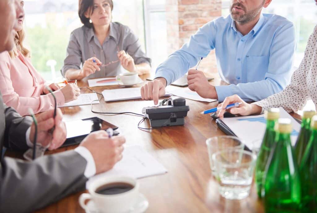 A group of business people sit around a wooden table in a meeting room, discussing the invoicing process while taking notes, with papers, tablets, water bottles, and a speakerphone in the center of the table.