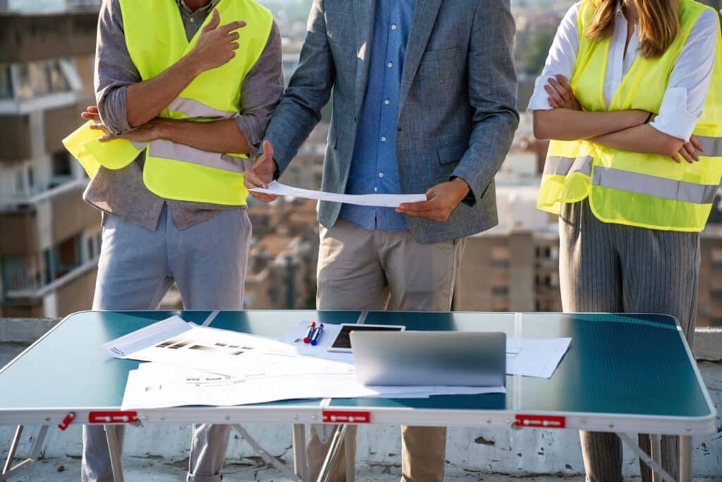 Three people in safety vests stand around a rooftop table, reviewing blueprints and documents with a laptop and papers—an essential scene for construction companies monitoring project progress, wip reporting, and financial health. Urban buildings rise in the background.