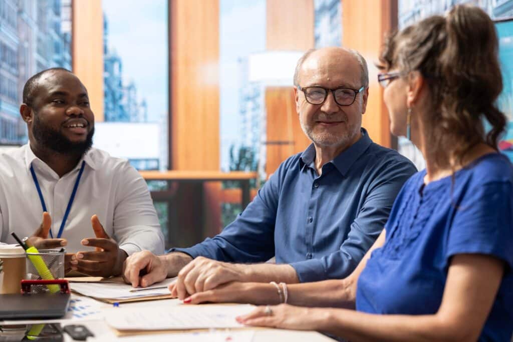 Three people are sitting at a table in an office, engaged in conversation about the account reconciliation process. One man is speaking and gesturing, while the others listen and smile. Papers and office supplies are on the table.