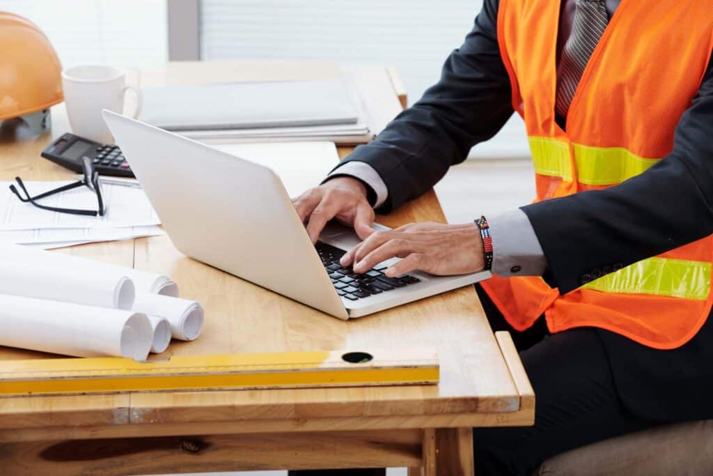A person wearing a suit and orange safety vest types on a laptop at a desk with blueprints, a level, documents, a hard hat, and a calculator—showcasing wip reporting for construction companies to track their financial health.