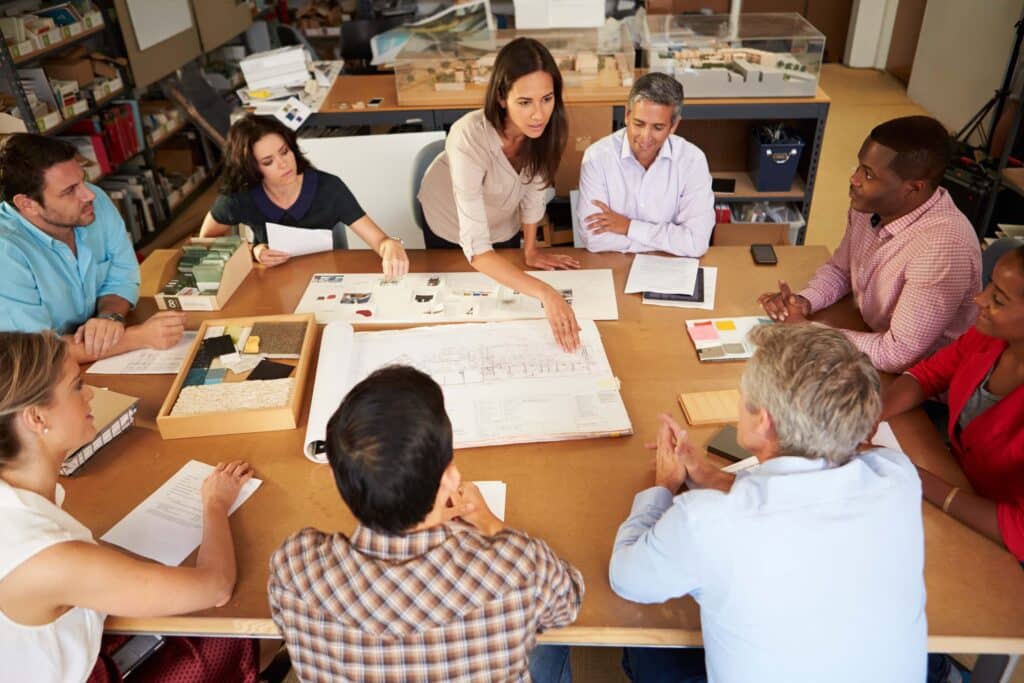 A group of eight people sits around a table in an office, discussing blueprints and documents. One woman stands and points to a plan, leading the project management meeting. Various materials and papers are spread out on the table.