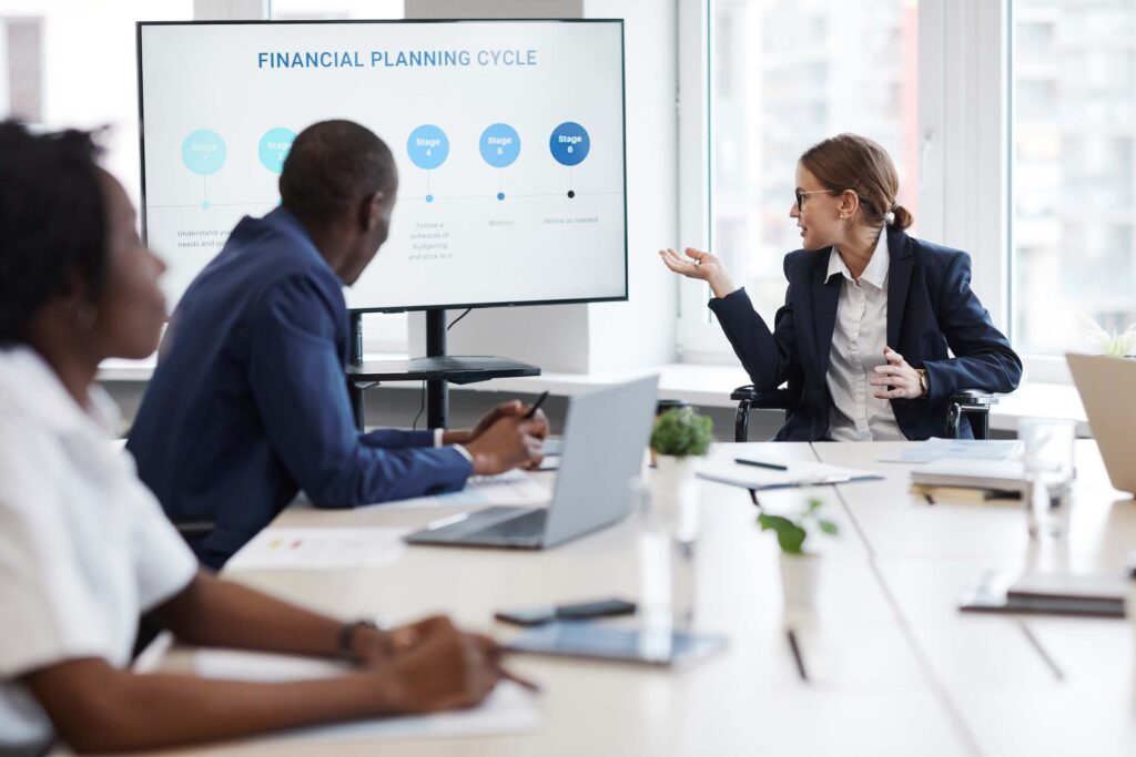 A woman in a suit presents a project financial management cycle chart on a screen to three colleagues in a bright conference room, with laptops, notebooks, and plants on the table.