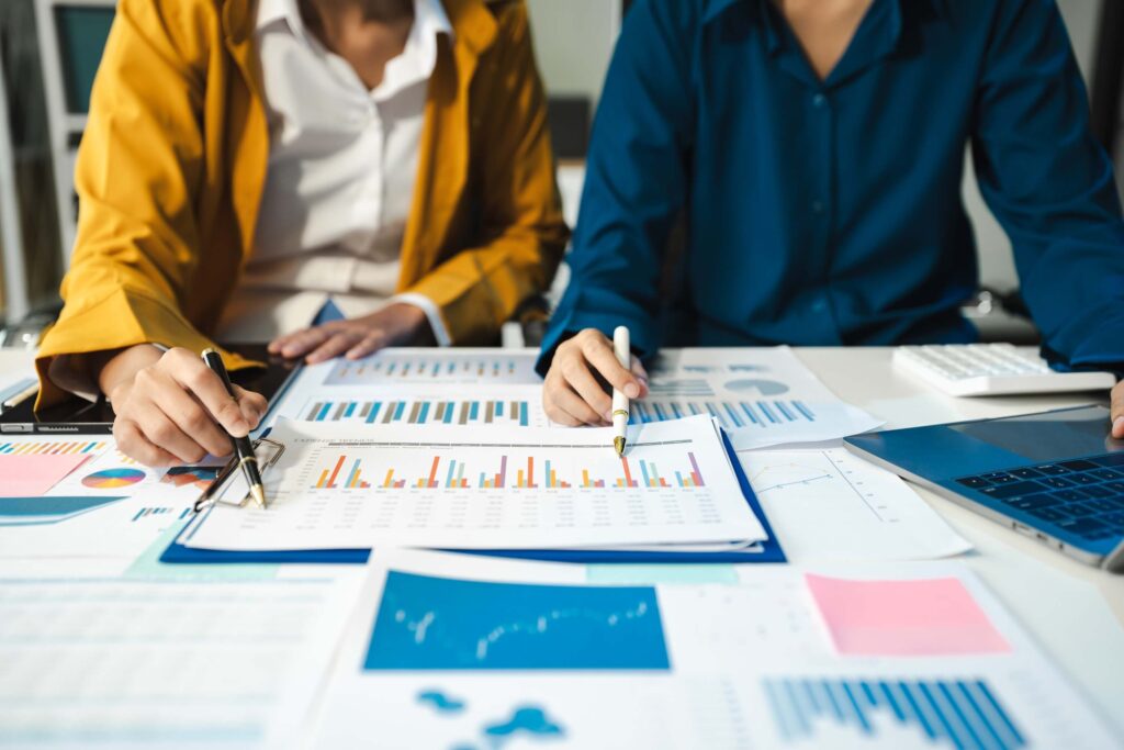 Two people in business attire analyze graphs and charts on paper at a desk, using pens to point out data related to wip reporting, with a laptop and various financial documents spread out in front of them.