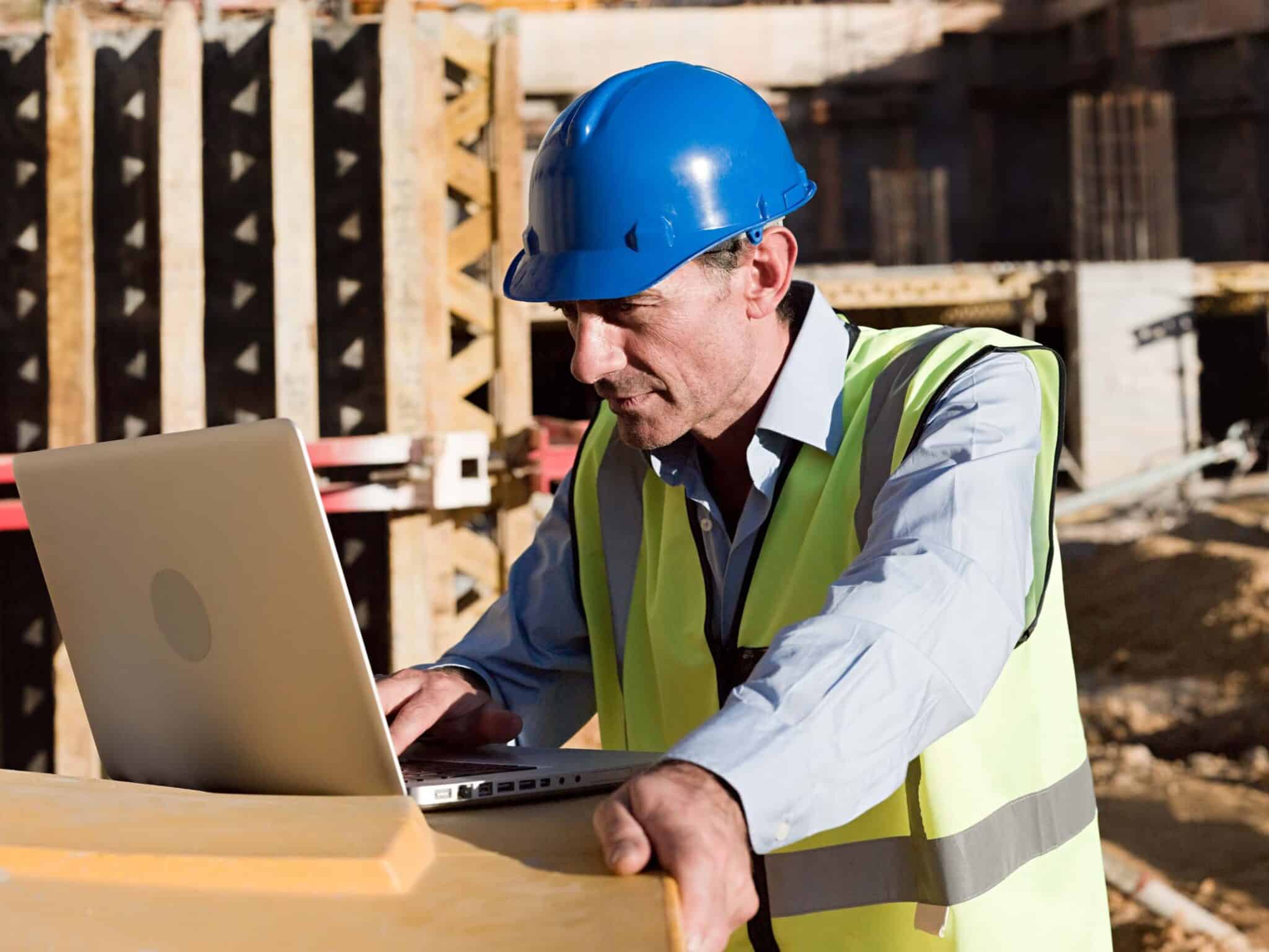 A construction worker wearing a blue hard hat and yellow safety vest uses a laptop at a construction site, efficiently managing change order tracking amid unfinished structures and building materials in the background.