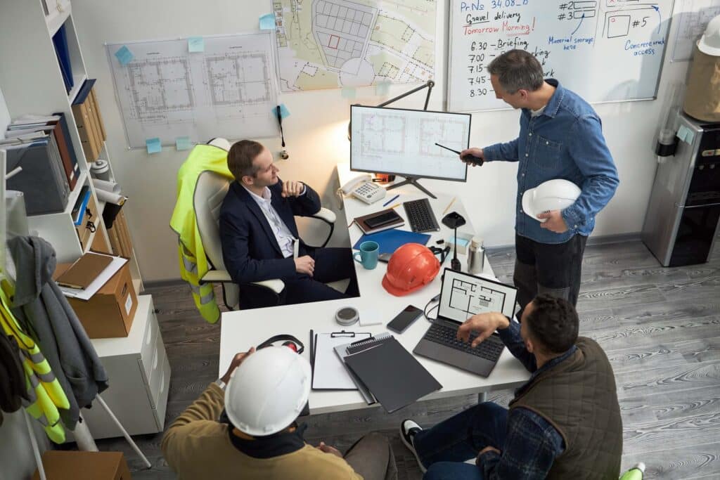 Four men in an office discuss architectural plans displayed on a monitor and paper. Two wear hard hats, one holds a white helmet, and construction gear is visible. A whiteboard with notes highlights change order tracking for project management.