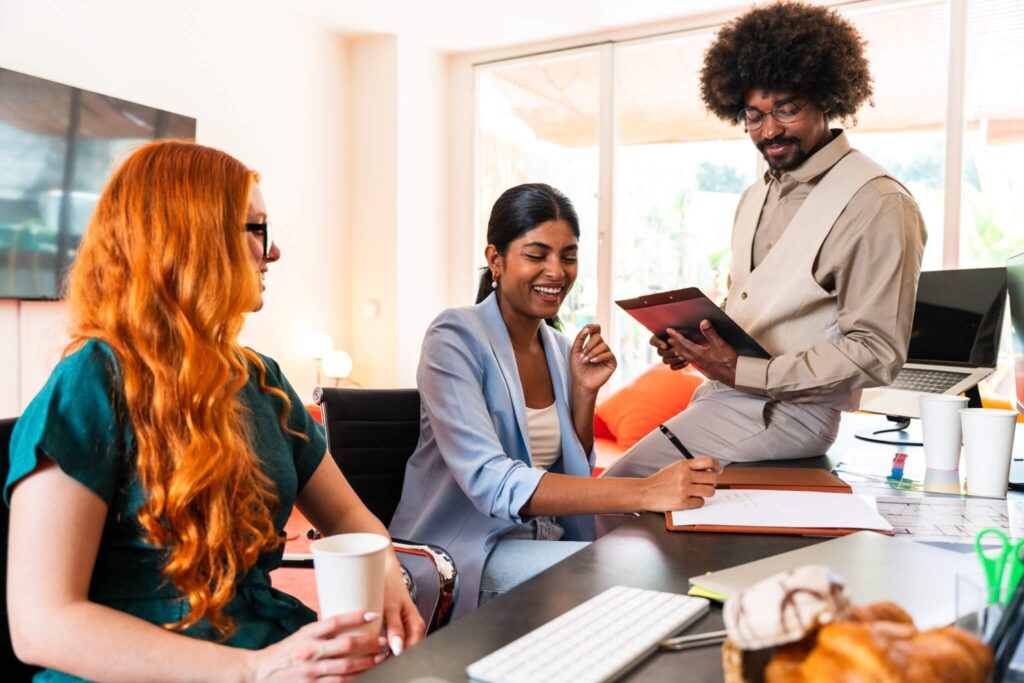 Three colleagues gather in a bright office. A woman with red hair holds a cup, another smiles while writing about the invoicing process, and a man with curly hair reads from a tablet while sitting on a desk.