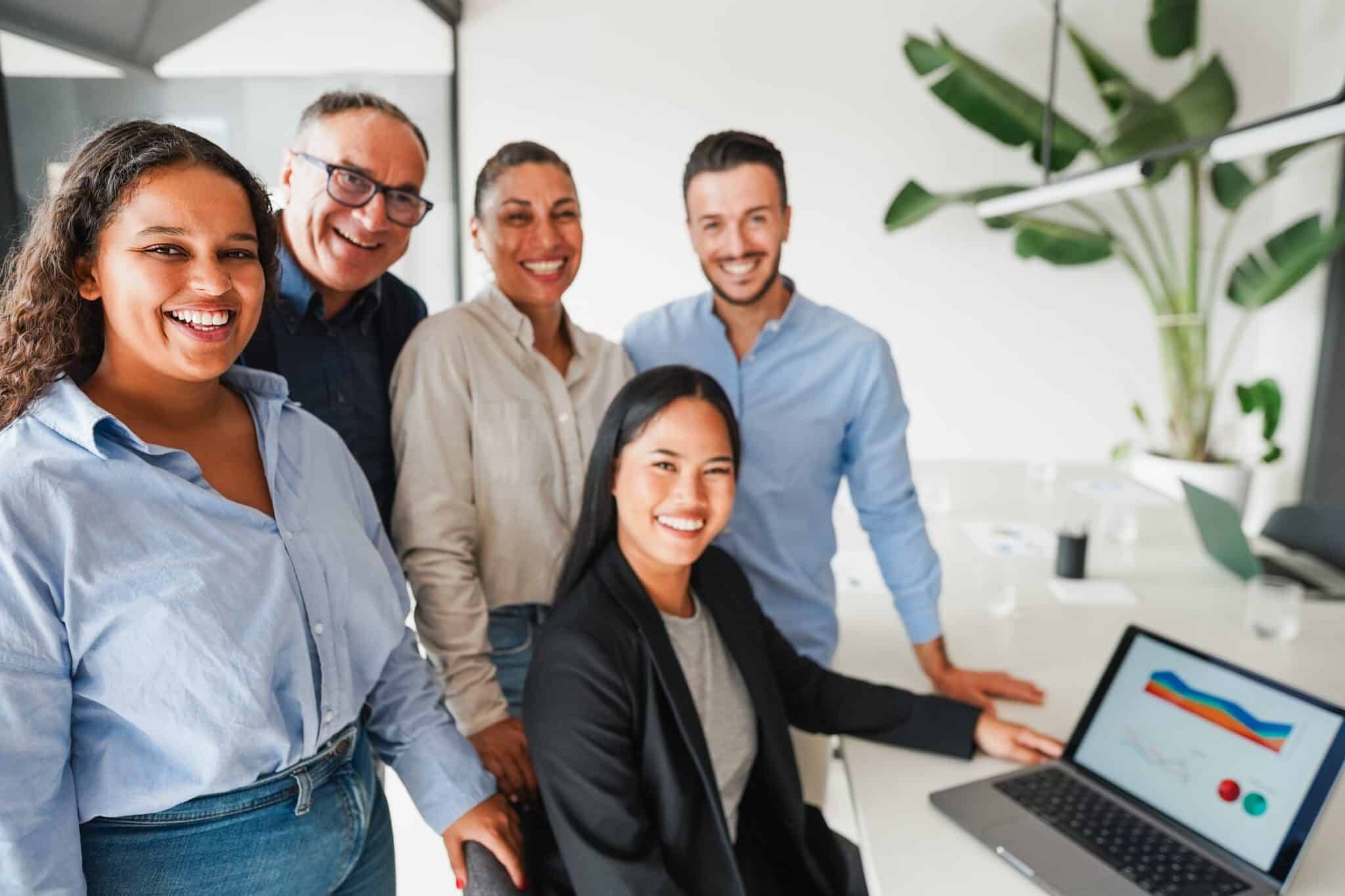 A group of five smiling people in business casual attire stand and sit around a table in a modern office, collaborating on the account reconciliation best practices. One person sits at a laptop displaying colorful charts. Large green plant in background.