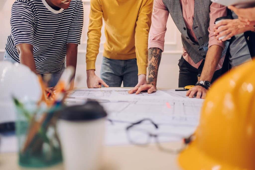 A group of people stand around a table, closely examining blueprints and documents; construction helmets, a coffee cup, and pens are visible in the foreground as they discuss change order tracking and the approval process.