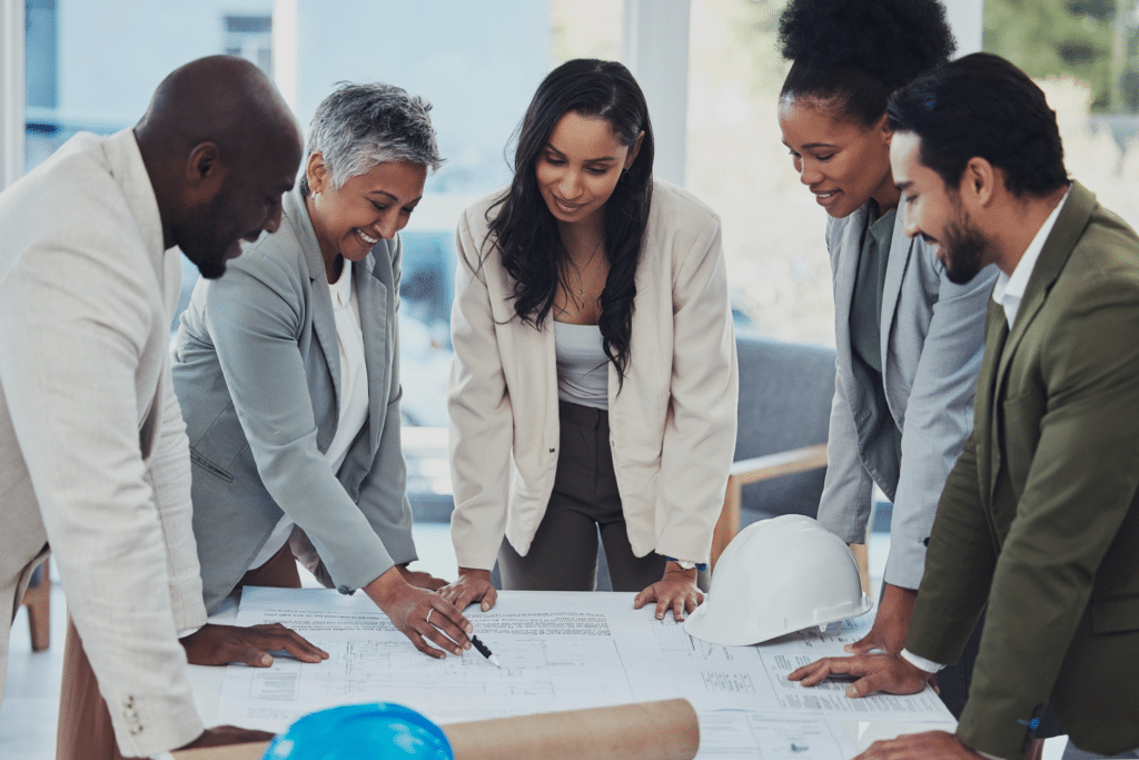 Five professionals, including project managers, stand around a table looking at blueprints and documents, collaborating in an office setting. Two safety helmets, one white and one blue, are visible on the table.
