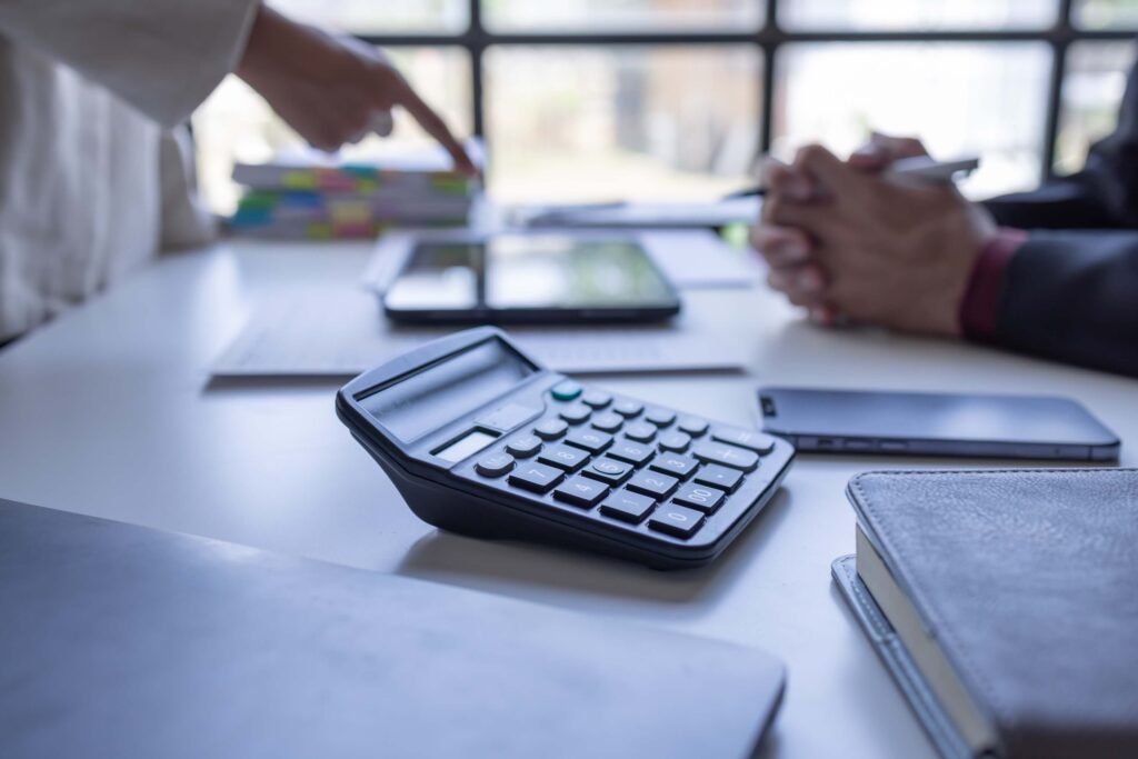 A close-up of a calculator on a desk with notebooks, a smartphone, and documents illustrates the account reconciliation process as two people discuss and point at a tablet in the background.