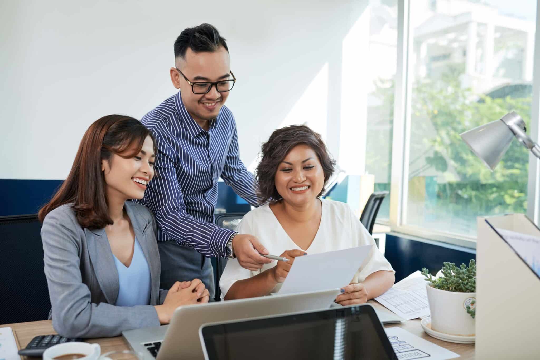 Three colleagues sit together at a desk, smiling and looking at a document about invoicing problems. One stands and points at the paper as they discuss invoicing problems, while the others sit with laptops and office supplies in a bright office.