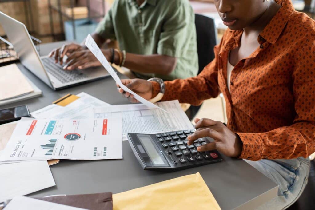 Two people working at a desk on the account reconciliation process with laptops, papers, charts, and a calculator. One uses the calculator and documents while the other types on a laptop amid financial documents and envelopes scattered around.