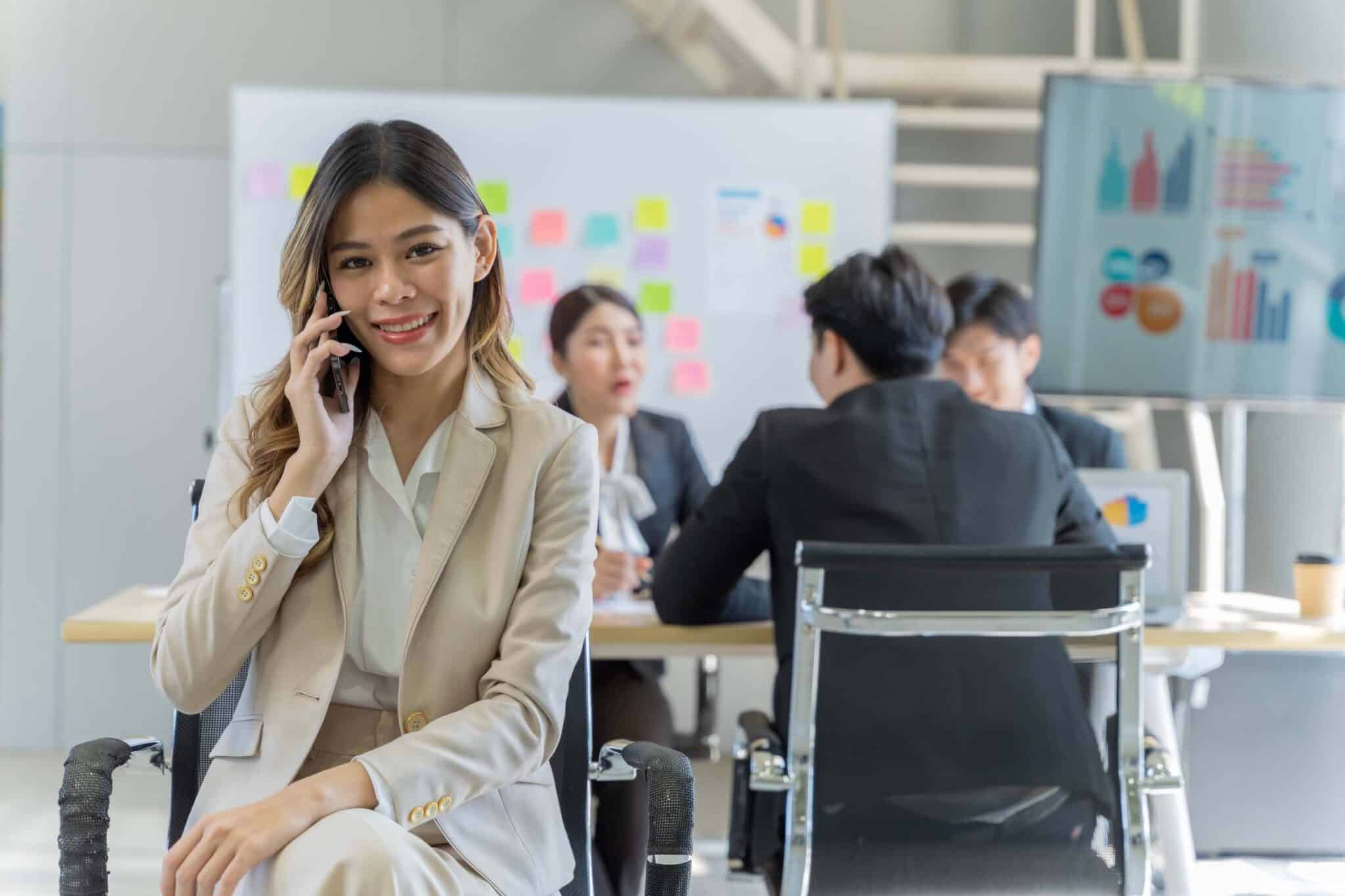 A woman in a beige suit smiles while talking on the phone in an office. Behind her, three colleagues have a meeting at a table with sticky notes and charts, discussing Deltek Replicon payroll time tracking software.