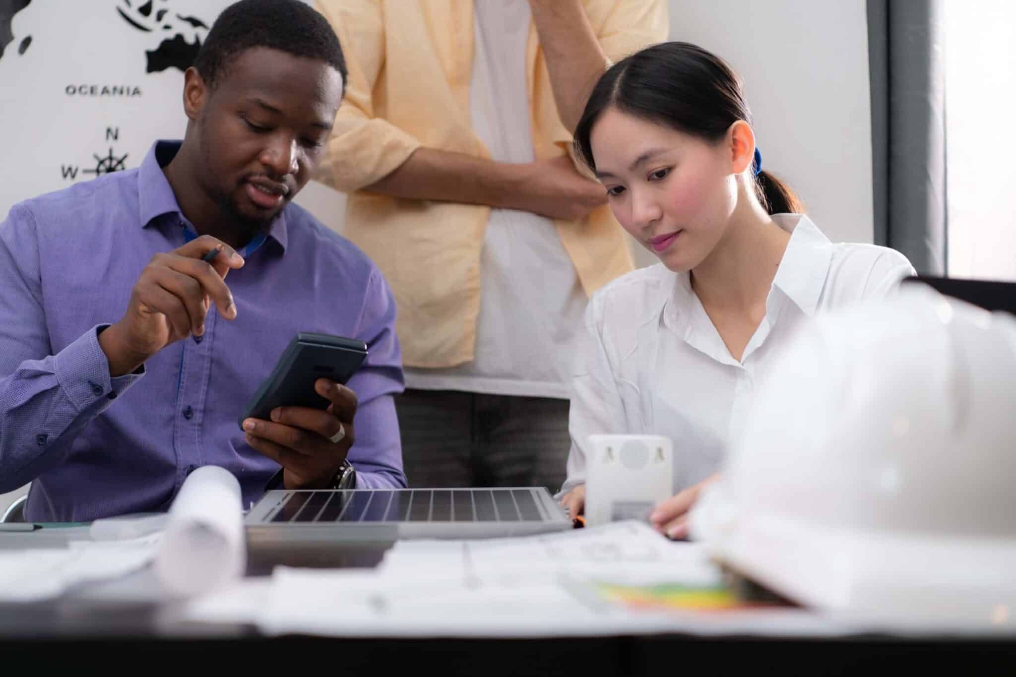 Two professionals sit at a desk covered with documents and a hard hat; the man holds a tablet displaying Replicon mobile time tracking software while the woman concentrates on her laptop. Another person stands in the background.