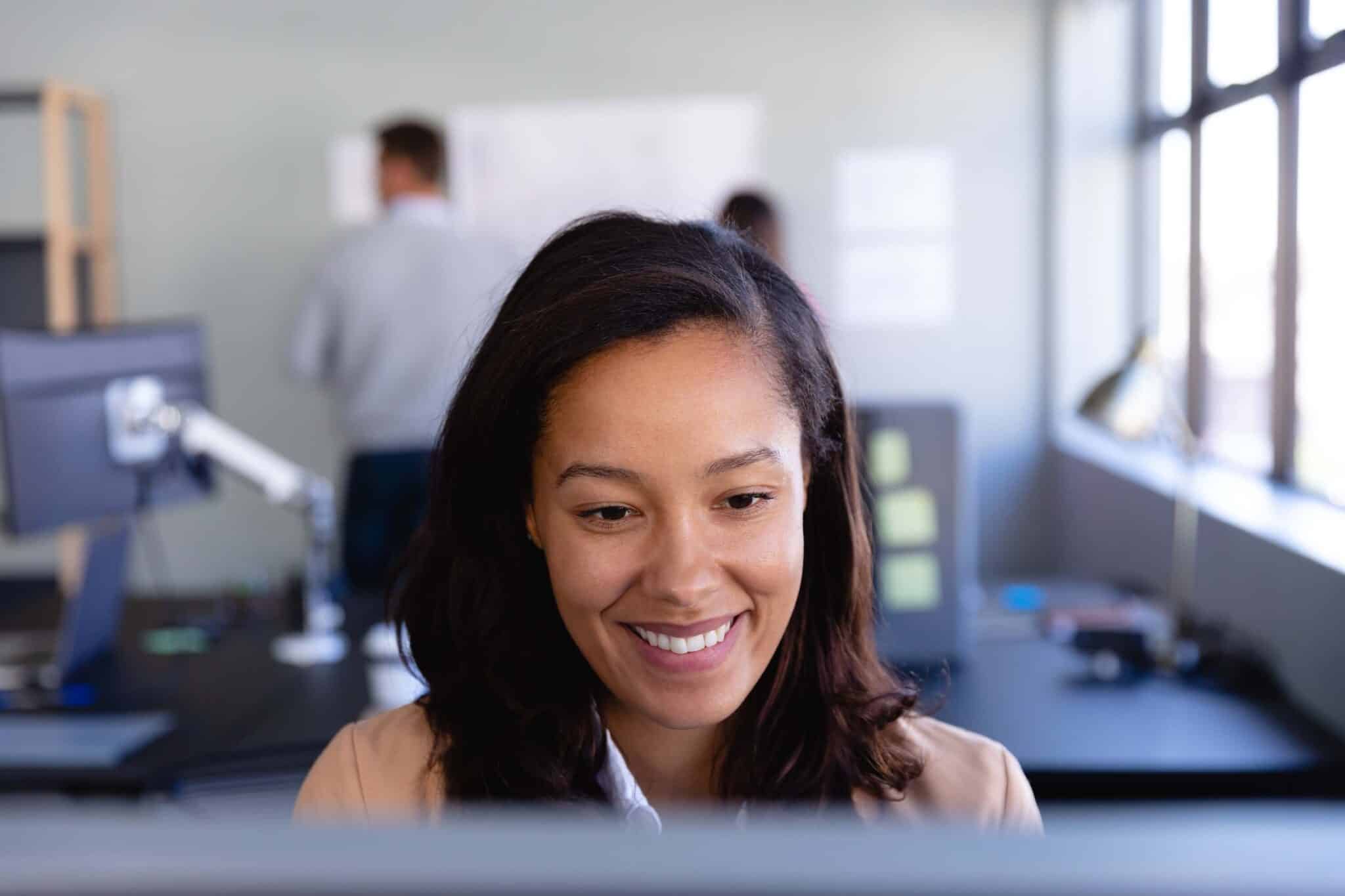 A woman with dark hair smiles while looking at a computer screen in a modern, bright office, possibly using Deltek Replicon payroll time tracking software. Two people are blurred in the background, working near desks and windows.