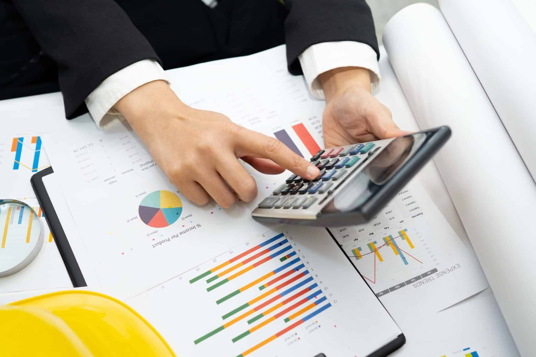 A person in business attire uses a calculator over documents with colorful charts and graphs, with a yellow hard hat, a magnifying glass, and Deltek Replicon payroll time tracking software on the desk.