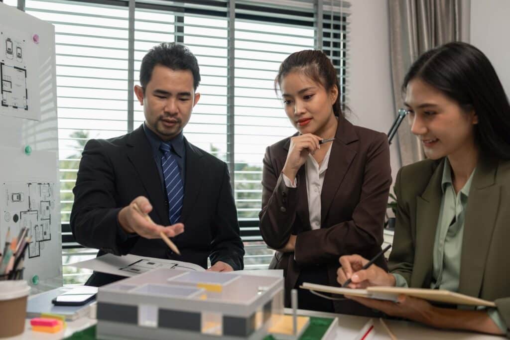 Three project managers in business attire discuss an architectural model at a bright office desk with plans and notes, appearing engaged and collaborative, perhaps reflecting on why most projects are still late despite thorough contingency planning.