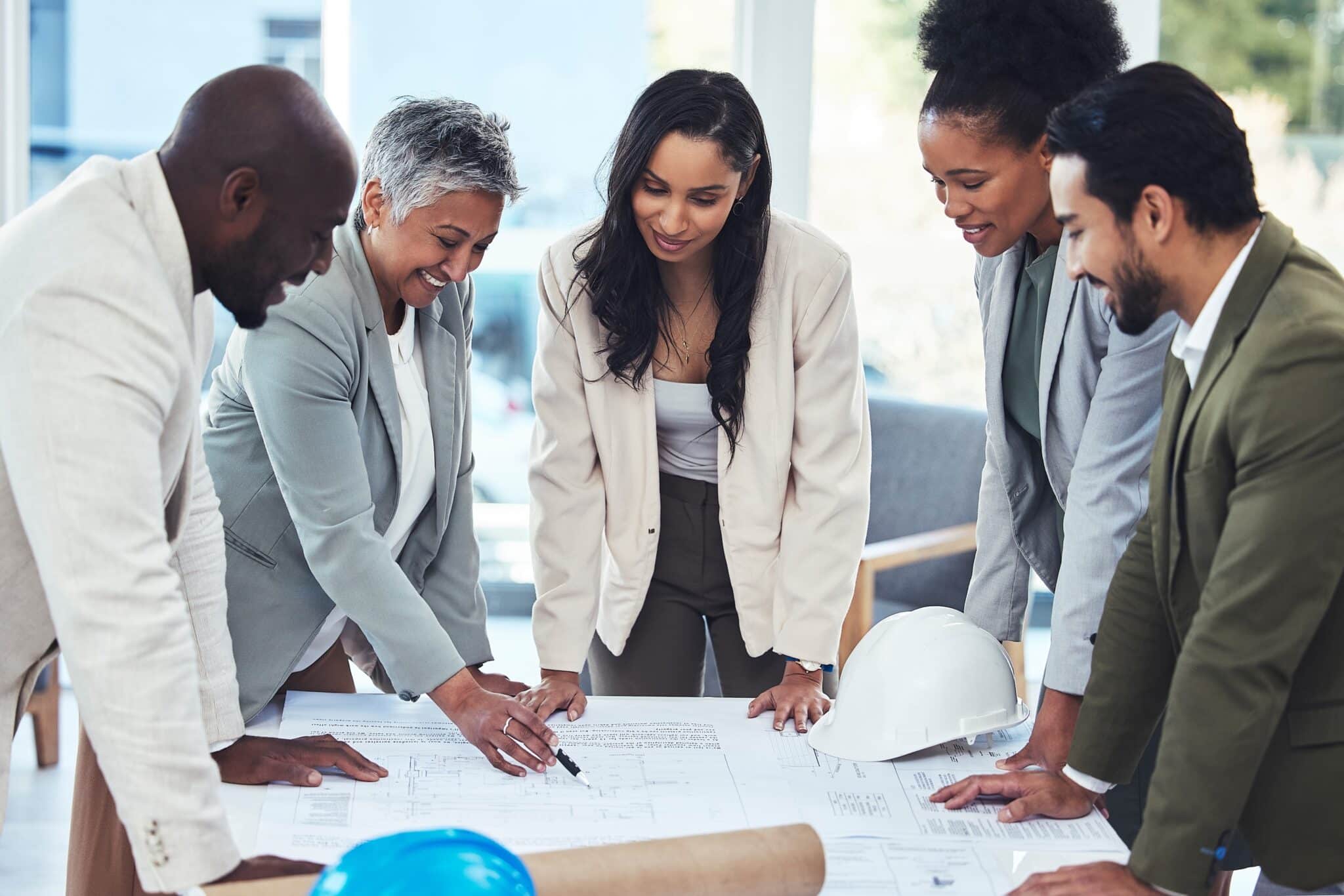 Five professionals in business attire stand around a table reviewing blueprints and documents, with Deltek Replicon payroll time tracking software visible on a laptop. A white and a blue hard hat suggest a collaborative planning or construction meeting.