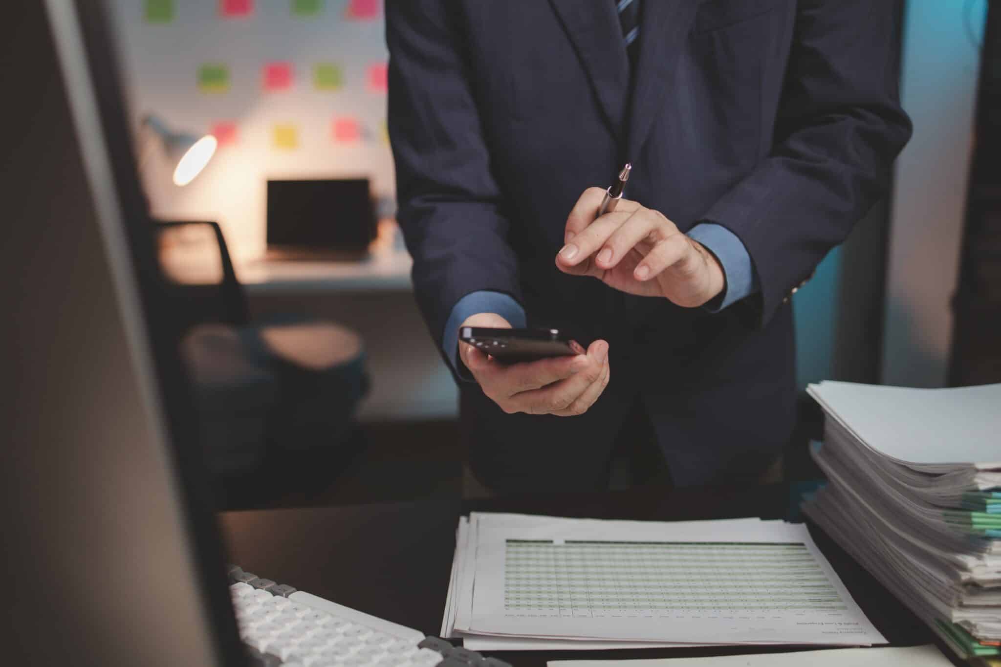 A person in a suit stands at a desk, holding a smartphone and a pen—perhaps using Replicon mobile time tracking software—as documents, a keyboard, and papers sit in the foreground; an office setting appears in the background.