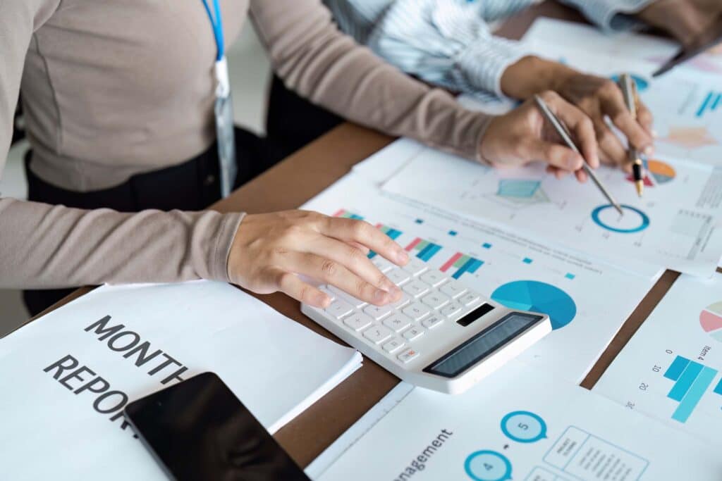 Two people from finance and accounting teams analyze financial statements and charts on paper; one uses a calculator, the other holds a pen. A "MONTHLY REPORT" document and a smartphone are also on the table.
