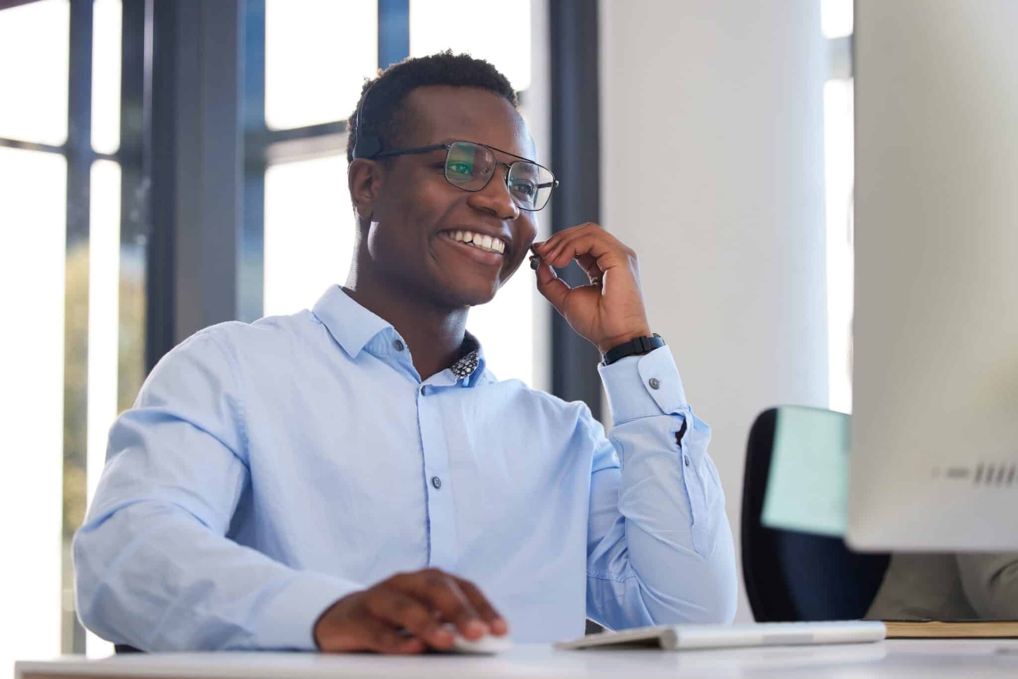 A man wearing glasses and a light blue shirt is smiling while talking on a headset and using Deltek Replicon payroll time tracking software at his desk in a bright, modern office.