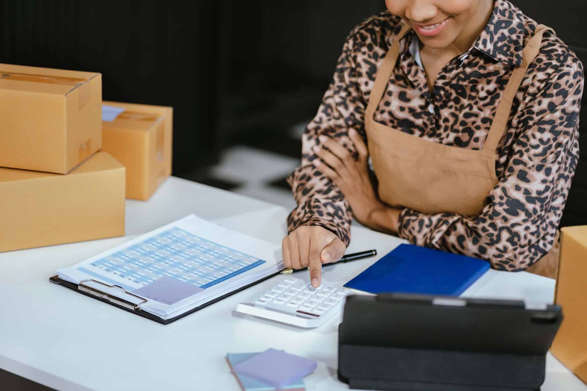 A woman in an apron sits at a desk with shipping boxes, charts, and a tablet. Using a calculator and wholesale accounting software, she smiles as she manages finances—capturing the spirit of small business or e-commerce success.
