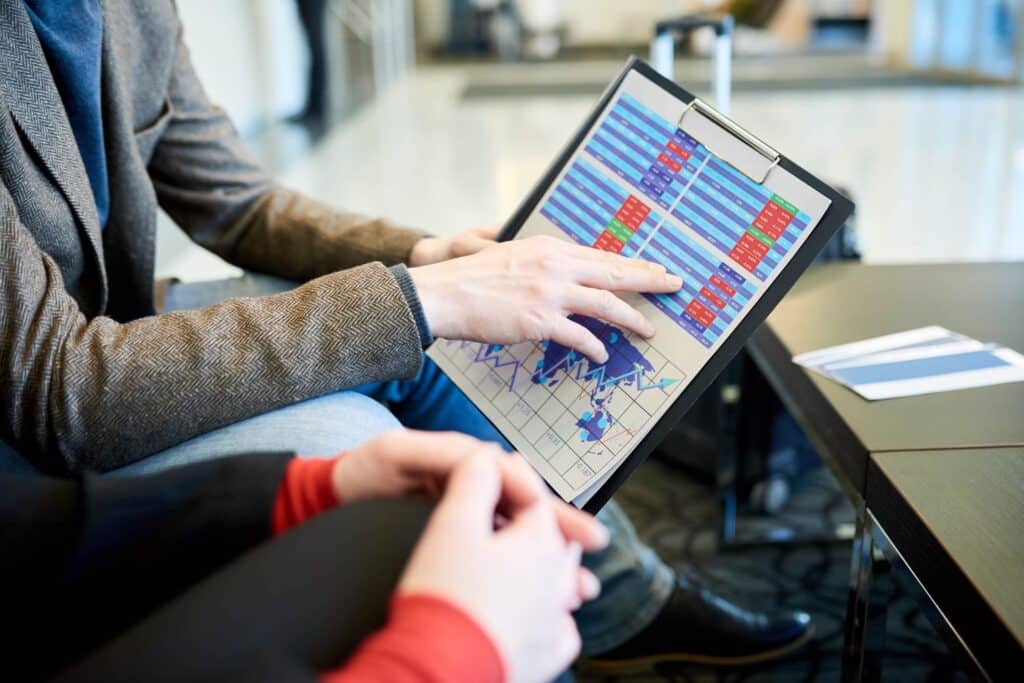 Two people sit together as one points to colorful charts and graphs on a clipboard, discussing cash flow forecasting and analyzing financial data in a modern office setting.