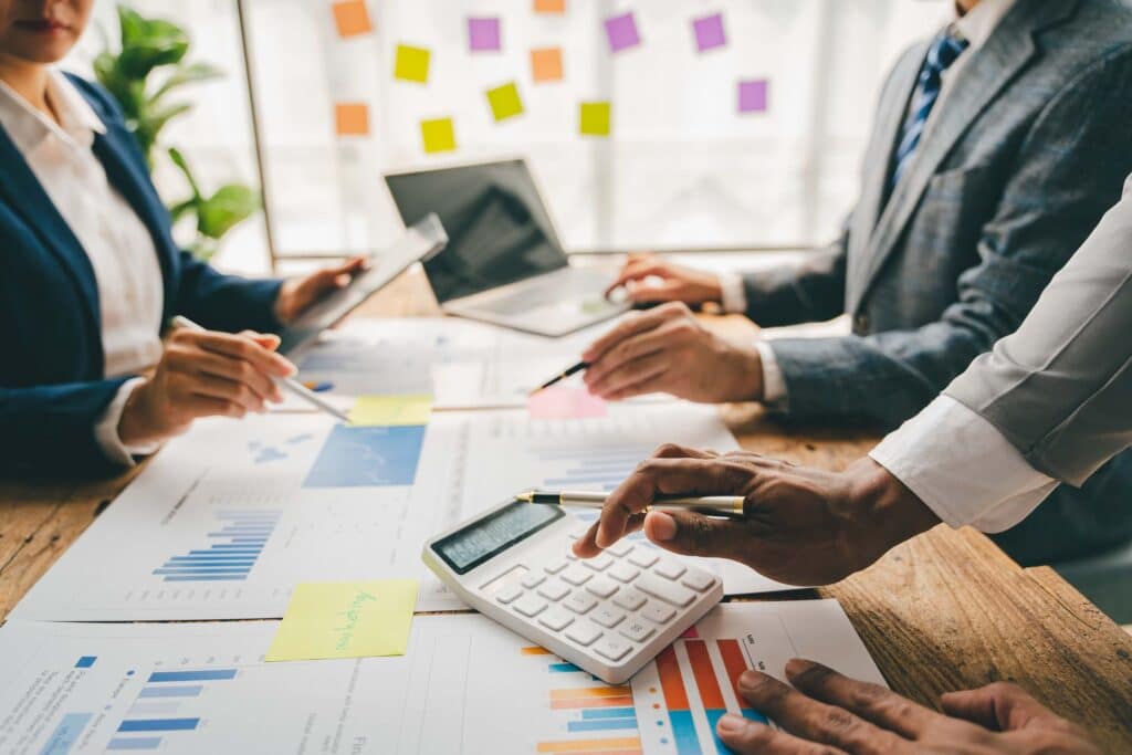 Three people in business attire review charts and graphs at a wooden table, using a calculator, tablet, and papers with financial data to analyze project margins. Colorful sticky notes and a laptop are visible in the background.