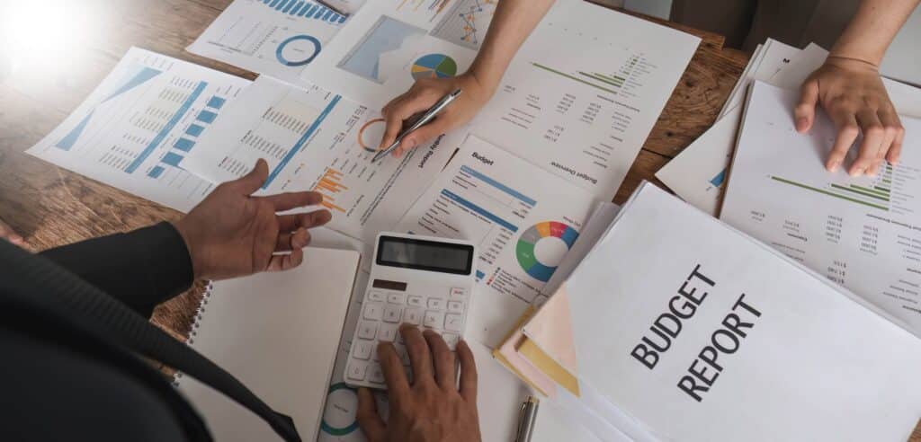 Three people are reviewing financial charts and graphs on a table with a budget report folder, calculator, and various documents displaying colorful data and pie charts for budget vs actual variance analysis.