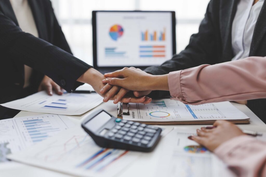 Four people in business attire put their hands together over a desk with charts, financial statements, and a calculator. In the background, a monitor displays colorful graphs and data, highlighting teamwork in finance and accounting teams.