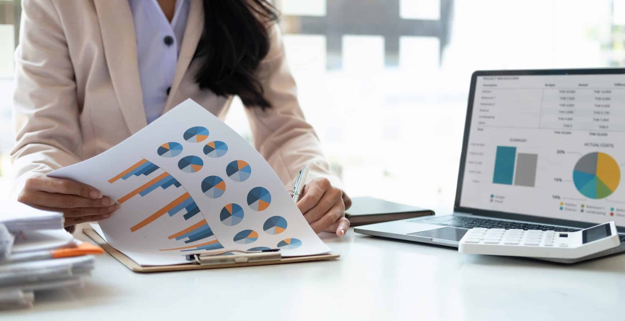 A person in business attire reviews printed charts and graphs at a desk with a laptop displaying Deltek Replicon payroll time tracking software, a calculator, and stacks of documents.