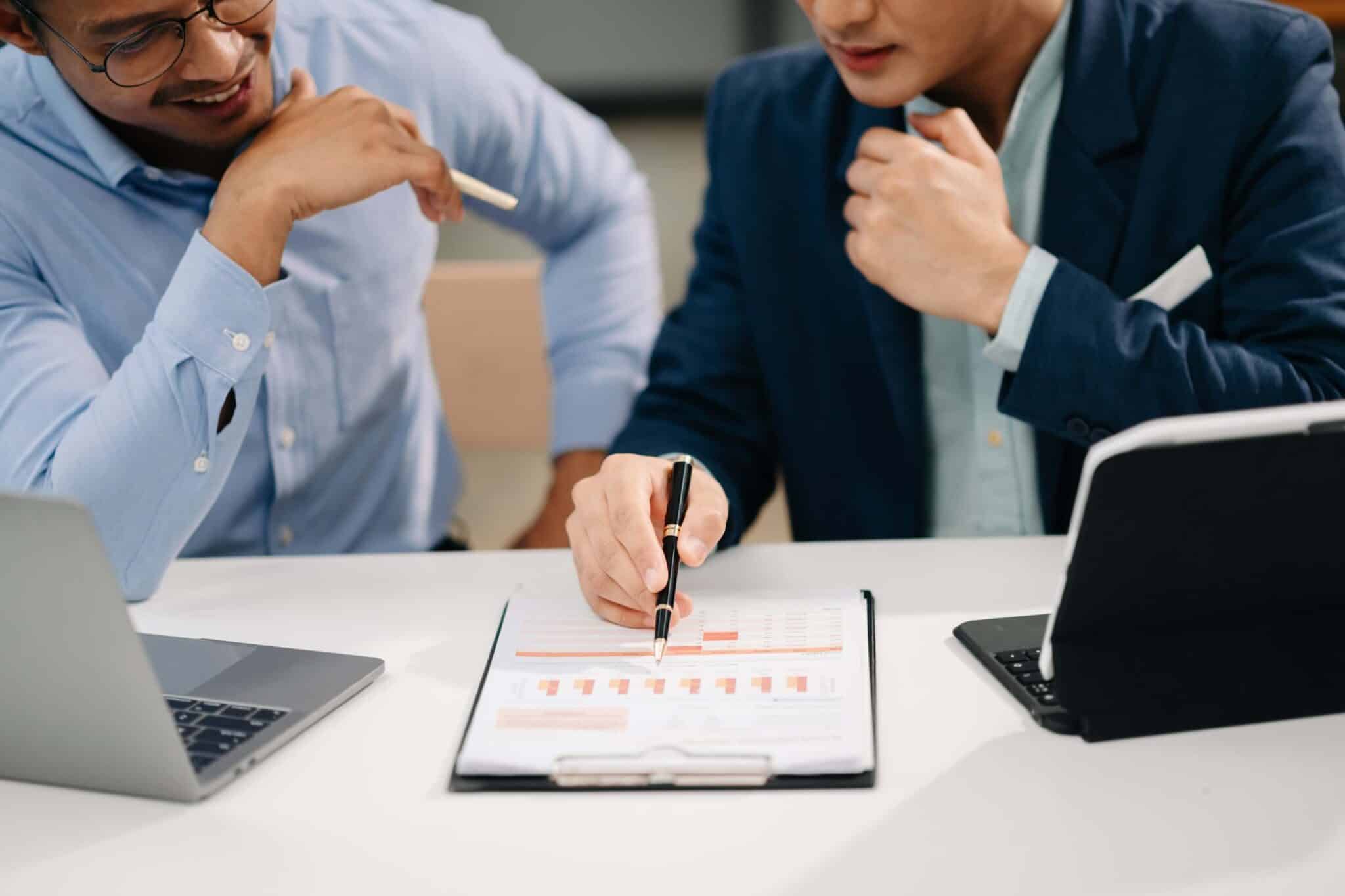 Two people in business attire sit at a desk with laptops, discussing a clipboard displaying charts and graphs from Replicon mobile time tracking software. One person points to the charts with a pen while the other listens attentively.
