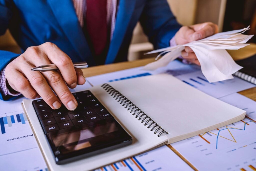 A person in a suit uses a calculator and holds several receipts, with an open notebook and financial charts on a desk, suggesting budget vs actual variance analysis as part of financial planning or accounting work.