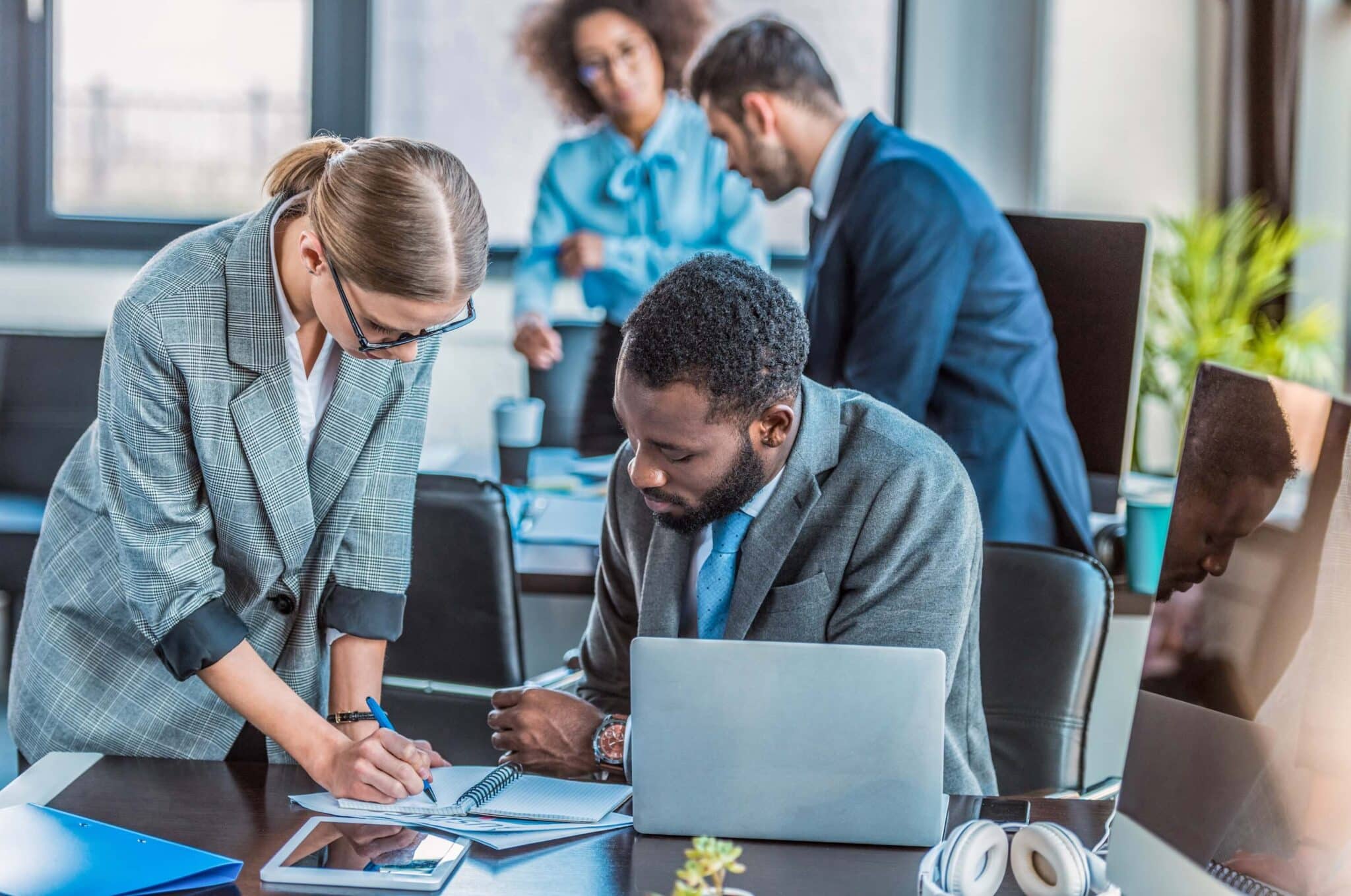Two business professionals collaborate at a desk with a laptop and documents, discussing Deltek Replicon payroll time tracking software, while two colleagues work together in the background in a modern office setting.