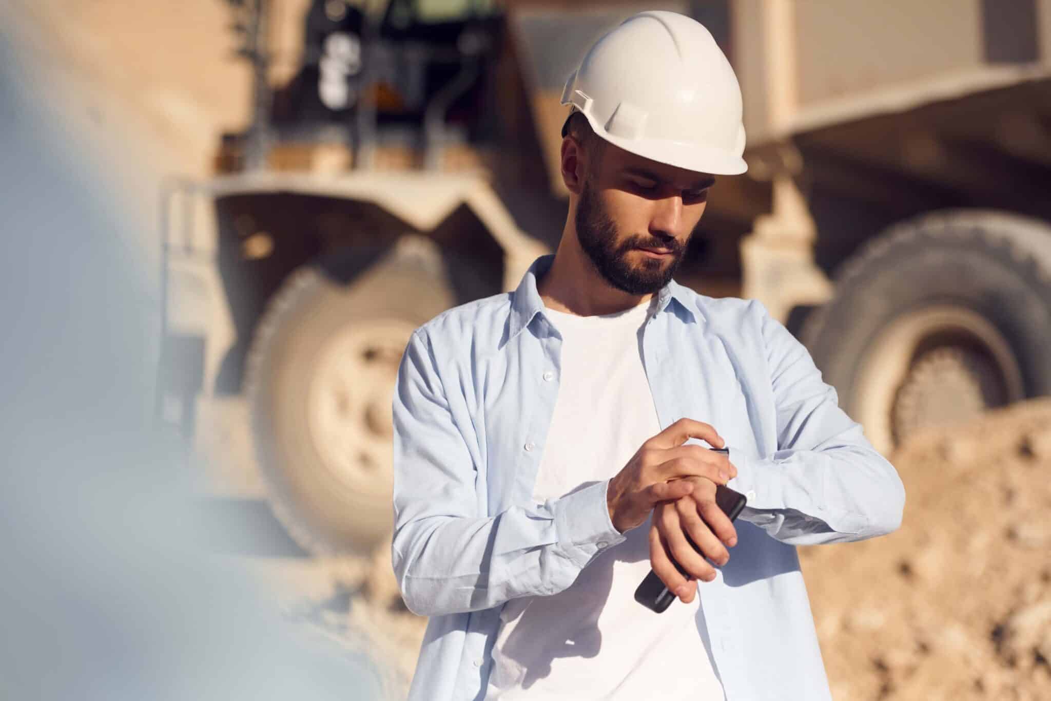 A man wearing a white safety helmet and light blue shirt stands at a construction site, looking down at his smartwatch, possibly checking Deltek Replicon payroll time tracking software, with large construction equipment blurred in the background.