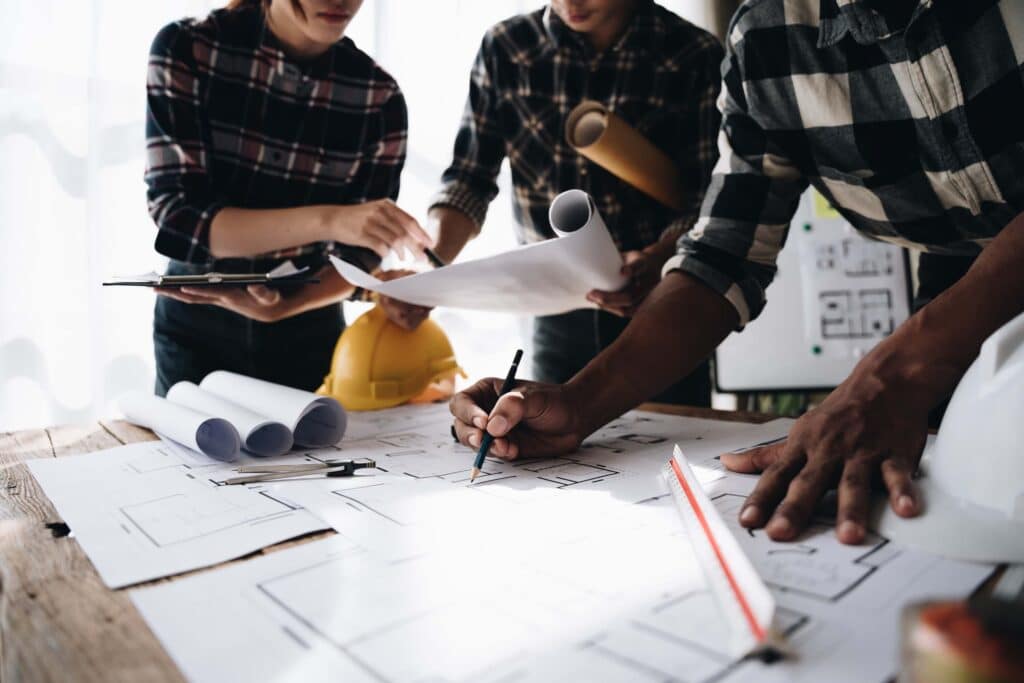 Three people review and discuss architectural blueprints on a table cluttered with rolled plans, papers, and a hard hat, debating project margins while one person draws on the plans and the others take notes. Project profitability is always a guess at this stage.