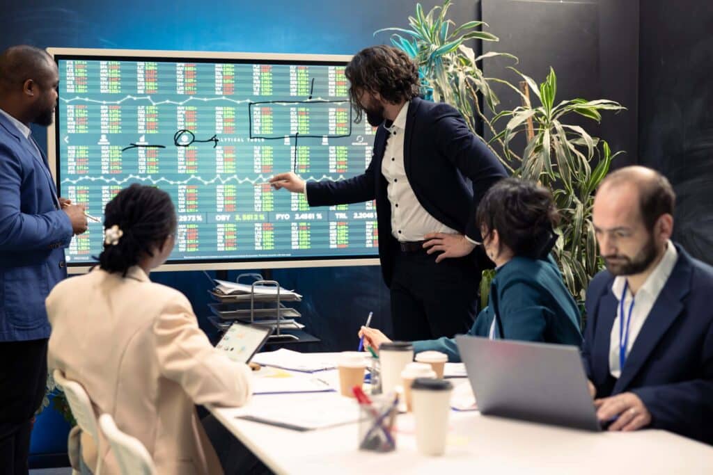 Four professionals in an office discuss project profitability shown on a large screen with charts and numbers, while two people sit at a table with laptops and documents, analyzing project margins for greater insight.