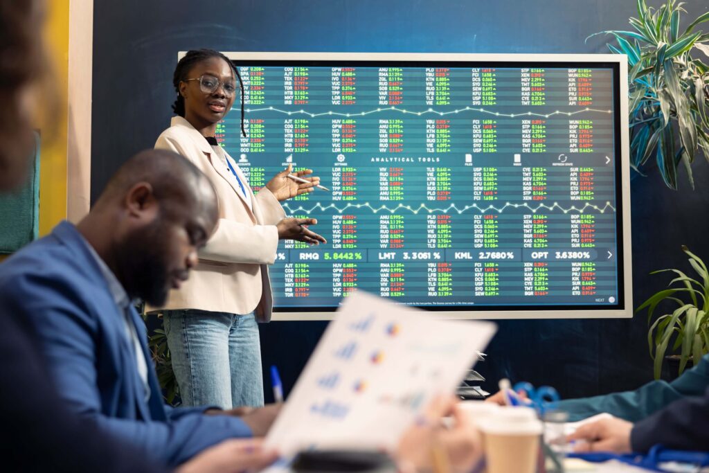 A woman stands by a large screen displaying colorful stock market data, pointing at it while presenting to a group of seated people focused on how to improve cash flow forecast with financial charts and notes.