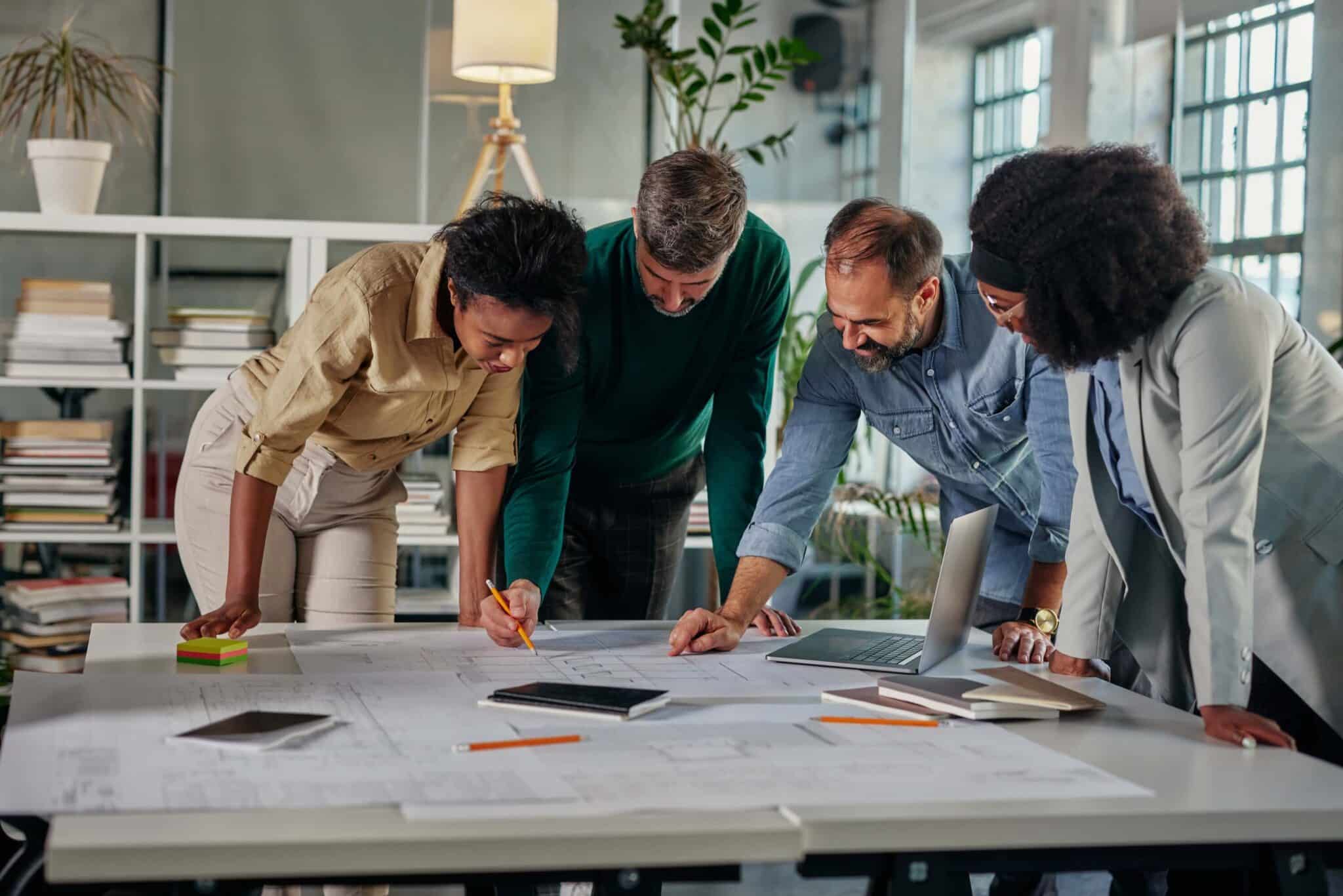 Four people stand around a table, collaborating and reviewing documents as they discuss how project profitability is always a guess. A laptop, notepads, and smartphones are on the table in the modern office with shelves, plants, and large windows.