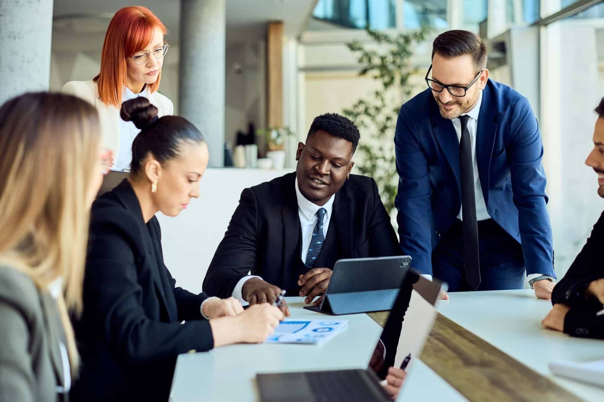 A diverse group of business professionals in formal attire collaborates around a table, reviewing Budget vs Actual Variance documents and digital devices in a bright, modern office setting.