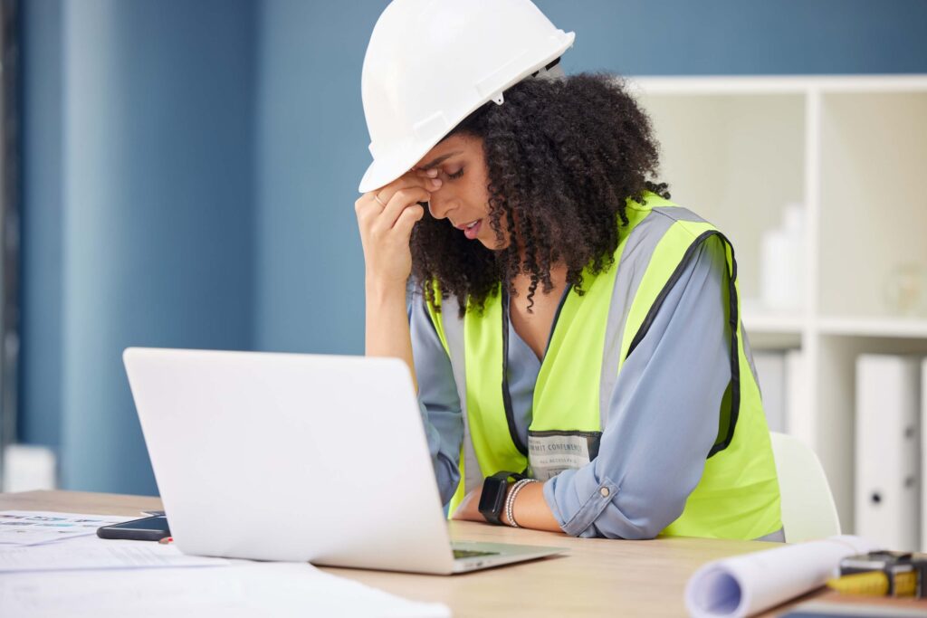 A woman wearing a hard hat and safety vest sits at a desk with a laptop, holding her forehead and appearing stressed—like many project managers who ask why are most projects still late despite contingency planning. Blueprints and a phone are on the desk in front of her.