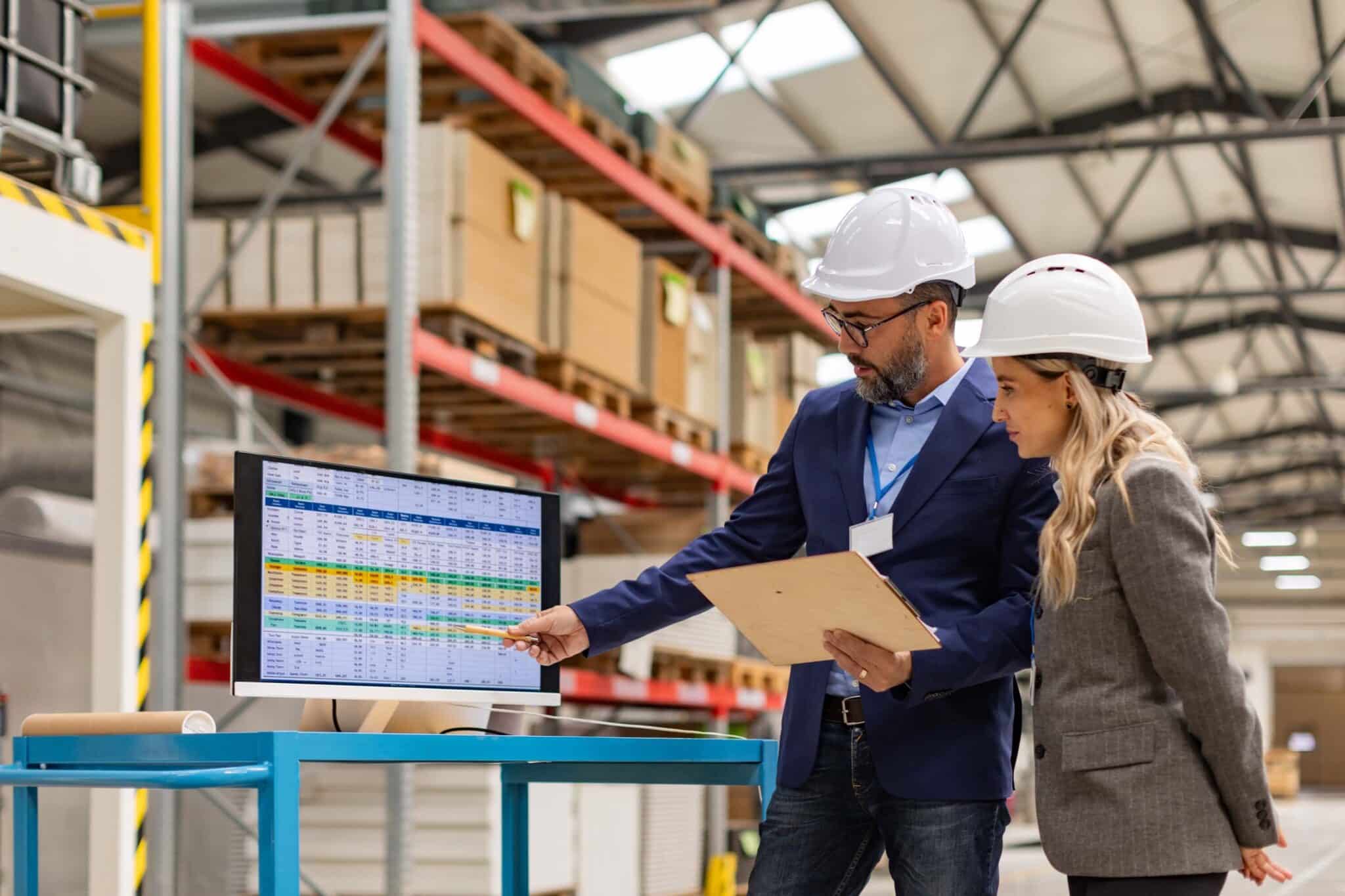 Two people in hard hats review data from wholesale accounting software on a computer screen in a warehouse. One points at the monitor while the other holds a clipboard, surrounded by shelves of boxes and industrial lighting.